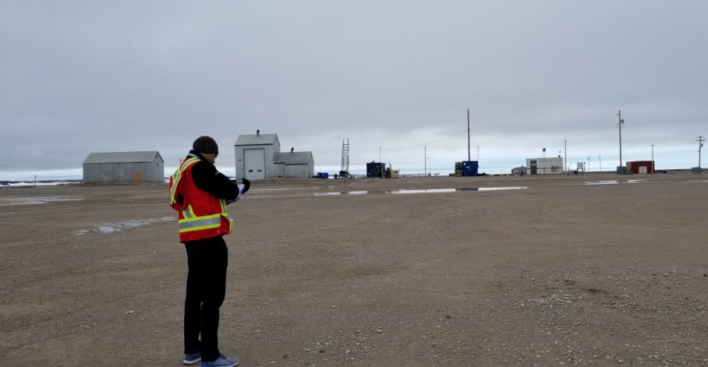 A WASCO employee is wearing a high visibility vest and writing on a clipboard, outside a small northern remote airport. Behind him is a large gravel lot with small buildings.